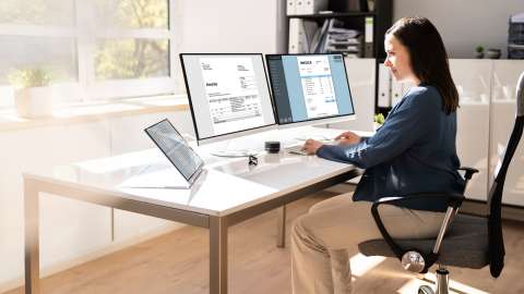 Um proprietário de uma pequena empresa está sentado em uma mesa dentro de um escritório moderno e cheio de luz revisando faturas exibidas em dois monitores de desktop lado a lado. Uma tabela exibindo uma planilha também está na mesa